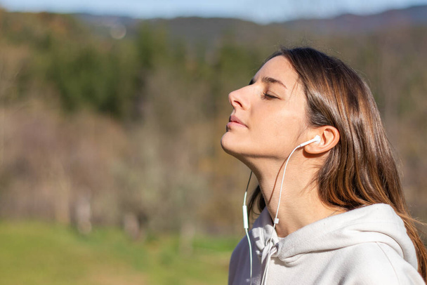 Mooie jonge vrouw ademt frisse lucht in het park luisterend naar muziek op koptelefoon. Ze draagt een lichte sweater en heeft lang bruin haar. - Foto, afbeelding