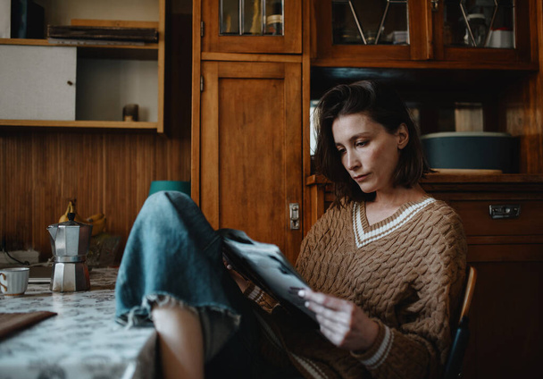Μια γυναίκα με πλεκτό μπεζ πουλόβερ και τζιν κάθεται στην κουζίνα και διαβάζει εφημερίδα.. - Φωτογραφία, εικόνα