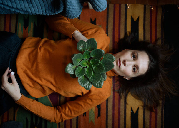 Ein Mädchen in orangefarbenem Pullover liegt auf einem bunten Teppich auf dem Boden und hält eine Zimmerblume auf der Brust - Foto, Bild