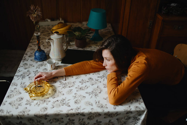 Una chica triste en un suéter marrón se sienta en la cocina con la cabeza apoyada en sus manos sobre la mesa - Foto, Imagen
