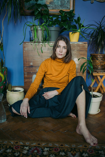 A young woman in an orange pullover and sea-green skirt sits on the living room floor surrounded by potted flowers - Foto, immagini