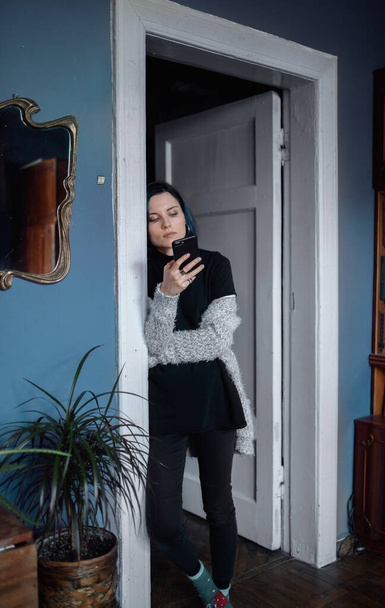 Una joven vestida de negro está de pie con un teléfono en las manos en la puerta de una sala de estar con paredes azules - Foto, Imagen
