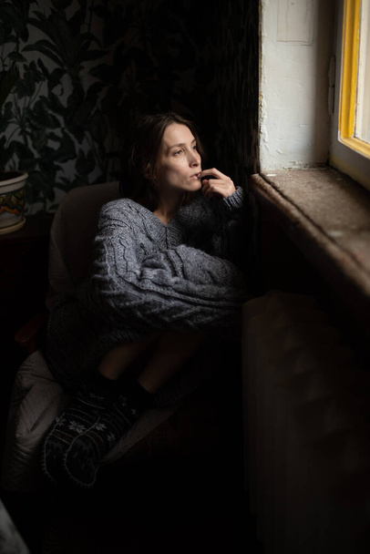 A pensive girl in a gray knitted sweater and warm socks sits on a chair with her legs crossed and looks out the window - Foto, Imagen