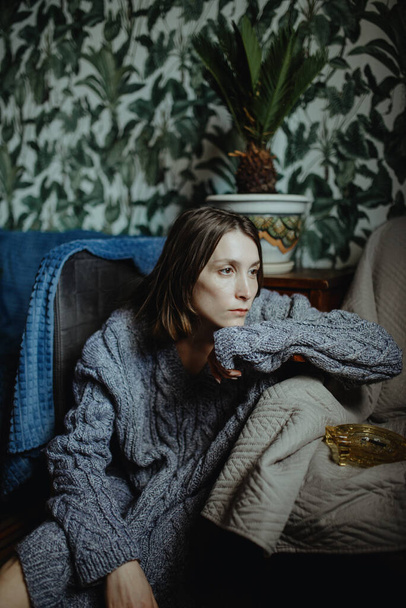 Fiatal szomorú lány szürke kötött pulóverben ül egy szobában egy széken csomagolással. - Fotó, kép