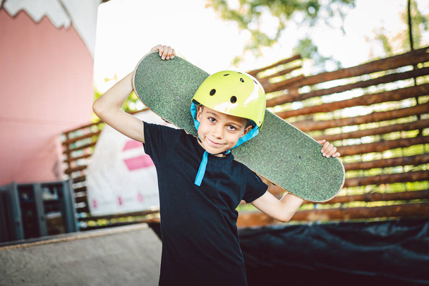 Портретный мальчик держит скейтборд через плечо. Красивая модель, позирующая на скейт-борде. Портрет стильный ребенок со скейт-бордом на полу трубы рампы открытый скейт-парк. Спорт, здоровье детей. - Фото, изображение