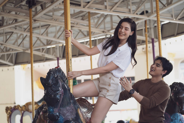 Young joyful couple man and woman riding on horse at Carousel amusement park. Concept happy and lovely life of teenager. - Photo, Image