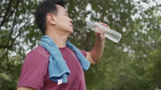 Asiático hombre mayor beber agua durante el descanso, mientras que el ejercicio en el parque con árboles en el fondo, de pie al aire libre refrescar relajarse después de hacer ejercicio, Feliz activo jubilado estilo de vida - Metraje, vídeo