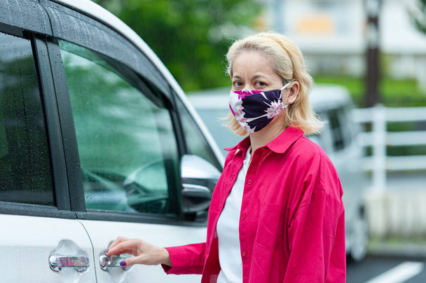 Salgın COVID19 - Coronavirus ve diğer bulaşıcı hastalıklara karşı korunmak için DIY ev yapımı bir maske takan karışık ırktan bir kadının portresi. Yağmurlu bir günde arabanın yanında.. - Fotoğraf, Görsel