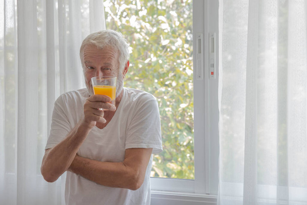 Senior retirement man stand beside room window holding a glass of orange juice look outside with happiness - Valokuva, kuva