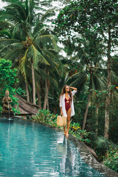 Woman in sunglasses, red swimsuit and white shirt relaxing and walking by poolside. Handmade straw bag. Infinity swimming pool in jungle. - Photo, Image