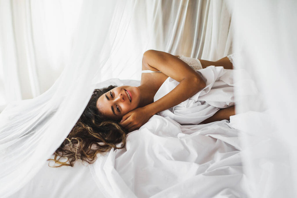 Beautiful tanned young woman awakening in white bed. Happy wake up and start new day. Leisure and rest. Wellbeing and carefree  concept. Long curly hair. Stay home. - Valokuva, kuva