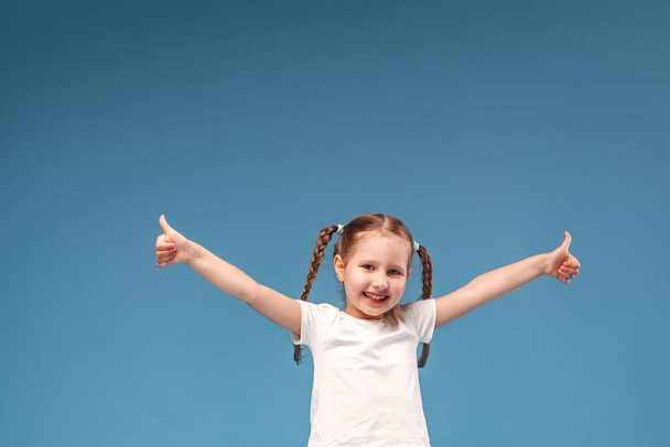 little girl with pigtails in a white t-shirt shows a gesture of approval to the class with both hands and is happy. Isolated on a blue background. - Fotoğraf, Görsel
