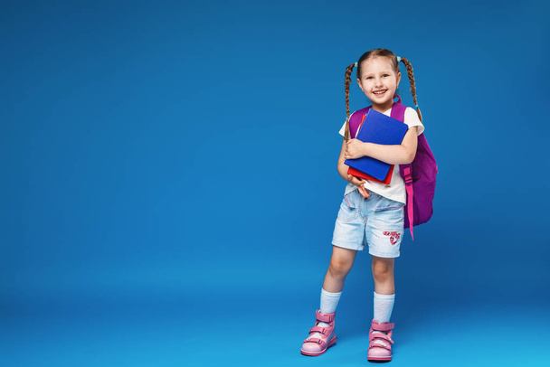 Happy smiling little girl goes to school for the first time. A full-length child with a school bag and a book in his hand on a blue background. Child in the classroom . Back to school. - Foto, Imagem