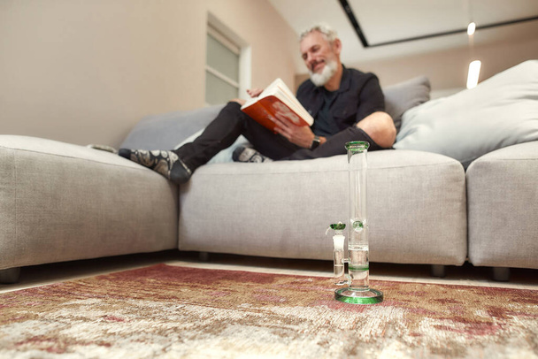 Tool for Relaxation. Bong or glass water pipe standing on the floor indoors. Bearded middle-aged man reading book while sitting on the couch in the background. Cannabis and weed legalization concept - 写真・画像