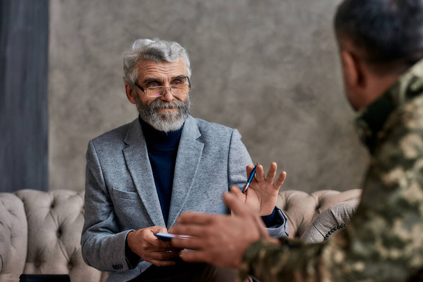 Getting back in shape. Portrait of mature psychologist communicating with military man and making notes during therapy session. Soldier suffering from depression, psychological trauma. PTSD concept - Fotografie, Obrázek