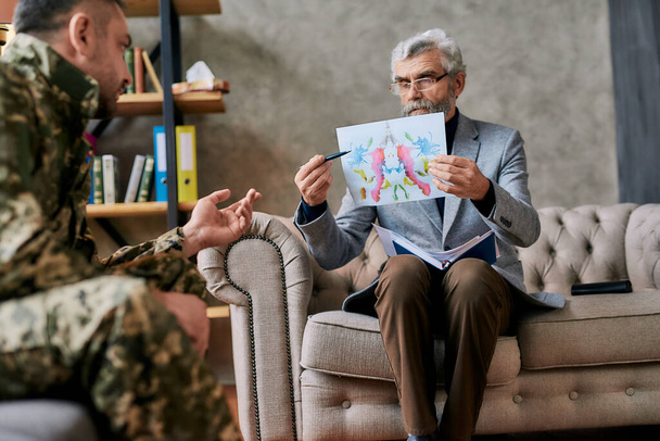 Mature psychologist holding picture with ink stain, Rorschach Inkblot in front of military man during therapy session. Soldier suffering from depression, psychological trauma. PTSD concept - Foto, Bild
