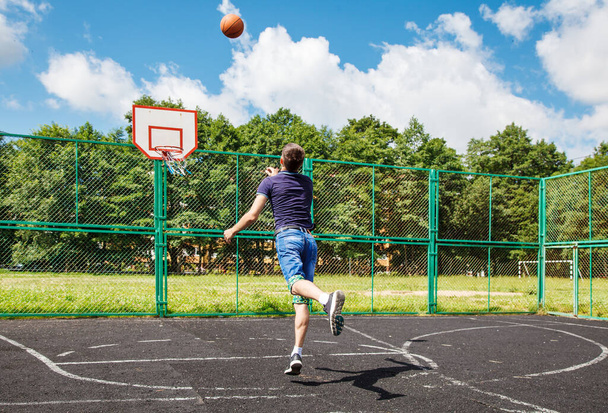 Młody mężczyzna w niebieskiej koszulce i jeansowych spodenkach grający w koszykówkę na boisku sportowym w słoneczny letni dzień - Zdjęcie, obraz