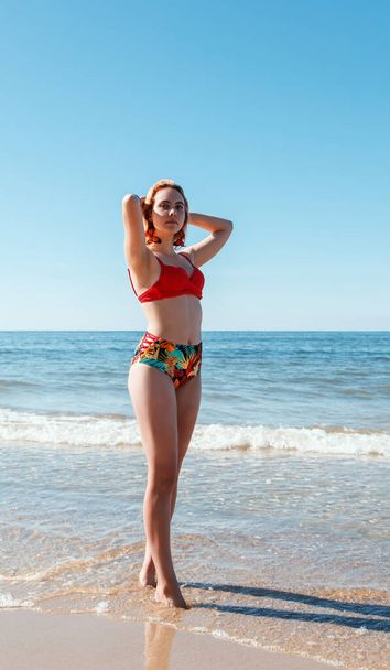 joven rubia en un traje de baño rojo de pie en la orilla del mar con las manos detrás de la cabeza en el soleado día de verano - Foto, imagen