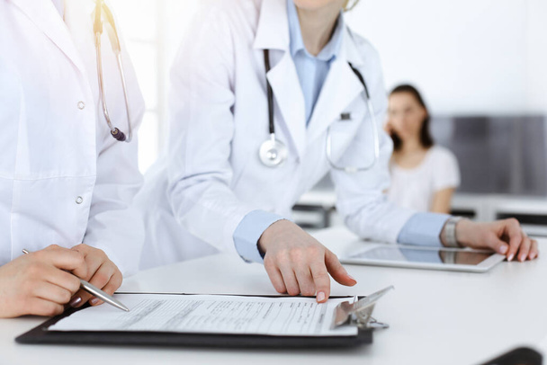 Unknown woman-doctors at work with patient at the background. Female physicians filling up medical documents or prescription while standing in hospital reception desk, close-up. Health care concept - Photo, Image