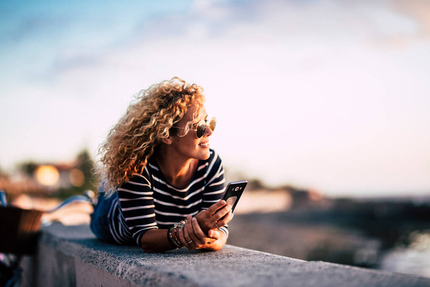 Beautiful middle age blonde curly woman enjoying and relaxing in outdoor leisure activity looking at sunset with modern phone ready to call friends  - Fotó, kép