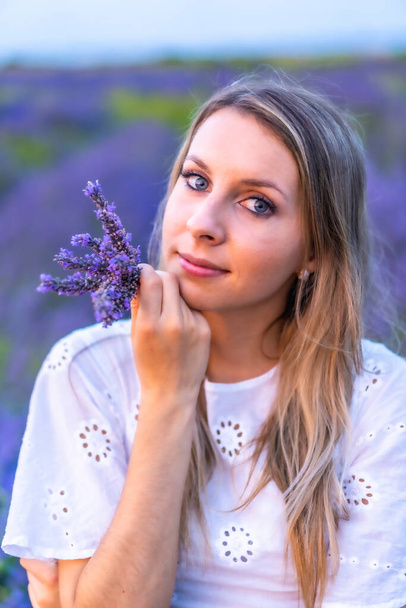 A young blonde Caucasian woman in a white dress in a cultivated lavender field in Navarra, Spain. lifestyle, rural lifestyle. Portrait with a bouquet of purple flowers - Photo, image