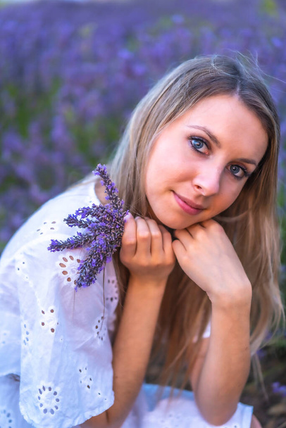 A young blonde Caucasian woman in a white dress in a cultivated lavender field in Navarra, Spain. lifestyle, rural lifestyle. With a bouquet of purple flowers - Photo, image