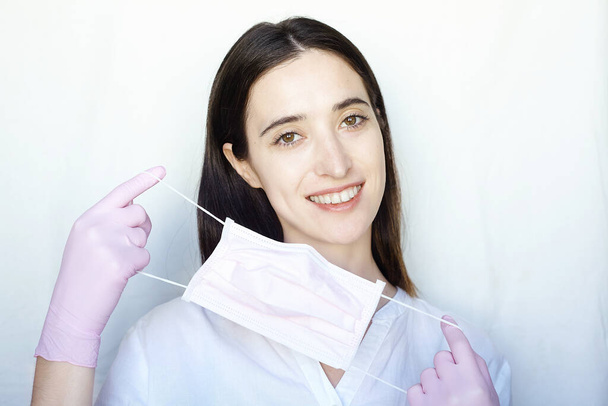 A woman in pink gloves puts on a pink protective mask. The girl stands on a light background in white clothes. The concept of suppression of coronavirus - Φωτογραφία, εικόνα