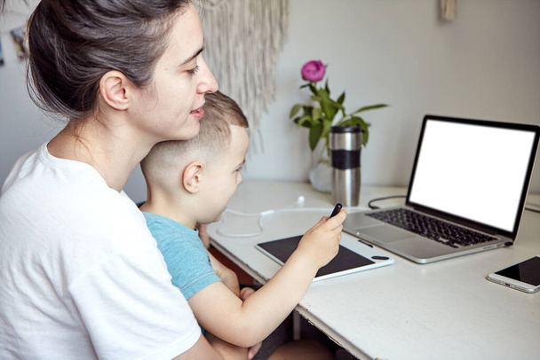 Eine Frau mit Kind arbeitet am Computer. Konzept der Arbeit von zu Hause aus und Familienerziehung. Mutter und Sohn arbeiten zu Hause am Grafik-Tablet und am Laptop. - Foto, Bild