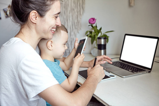 子供を持つ女性はコンピュータで働いています。家庭や家庭教育からの仕事の概念。ママと息子はグラフィックタブレットやノートパソコンで働いてる. - 写真・画像