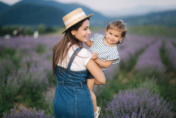 Portrait of young mom with her adorable daughter in summer lavender field. Family denim style. Woman in straw hat - Photo, image
