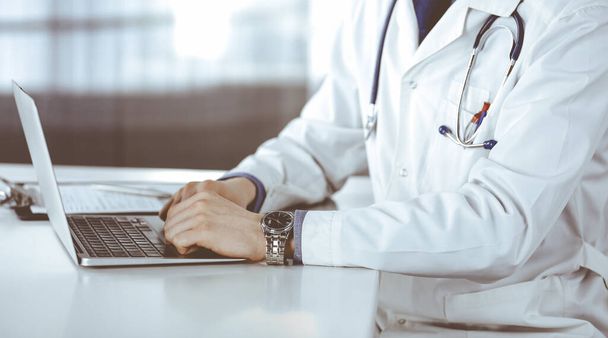 Unknown male doctor sitting and working with laptop in clinic at his working place, close-up. Young physician at work. Perfect medical service, medicine concept - 写真・画像