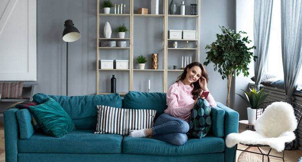 Beautiful smiling alone woman with smartphone on sofa in the interior of a living room. Use on mobile at home. - Foto, Imagem