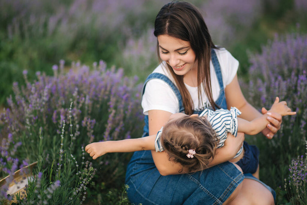 Porträt einer jungen Mutter mit ihrer entzückenden Tochter im sommerlichen Lavendelfeld. Familienfreundlicher Jeans-Stil. Frau mit Strohhut. Mutter kitzelt Tochter - Foto, Bild