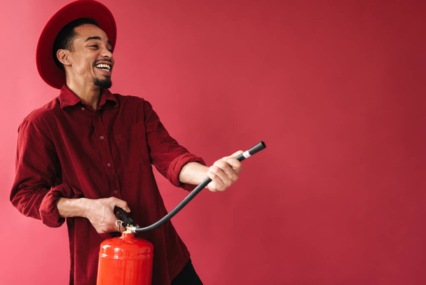 Cheerful smiling stylish young african man using fire extinguisher while standing isolated over red background - Foto, Imagem