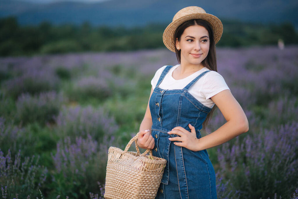Portrait of attractive woman in lavender field in straw hat and denim sundress. Farm style in lavender with straw bag - Zdjęcie, obraz