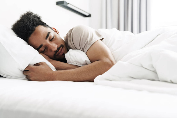 Photo of young african american man with mustache sleeping in white bed at bright room - Φωτογραφία, εικόνα