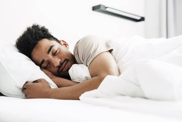 Photo of young african american man with mustache sleeping in white bed at bright room - Foto, Imagen