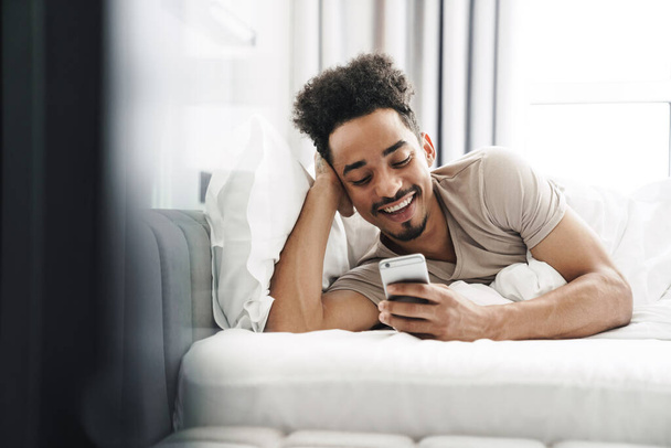 Photo of happy african american man with mustache using mobile phone and smiling in bed at bright room - Foto, afbeelding