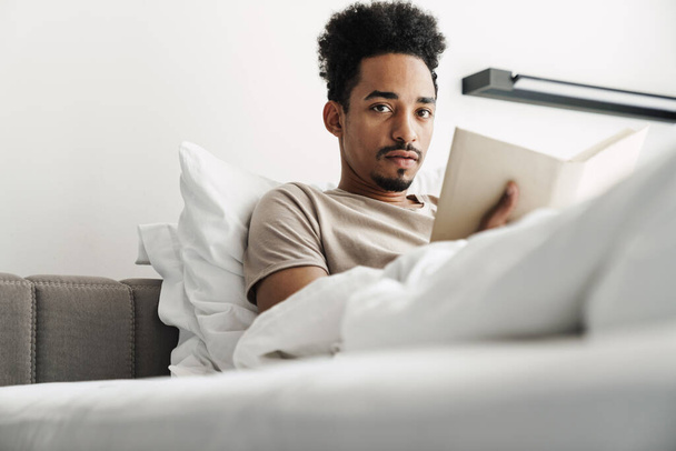 Photo of serious african american man with mustache reading book while lying in bed at bright room - Photo, Image