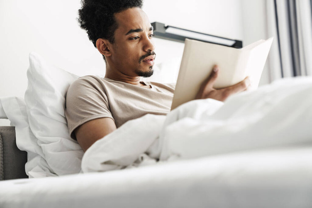 Photo of serious african american man with mustache reading book while lying in bed at bright room - 写真・画像