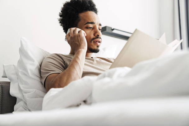 Zdjęcie poważnego Afroamerykanina z wąsami czytającego książkę podczas rozmowy przez telefon komórkowy w łóżku w jasnym pokoju - Zdjęcie, obraz