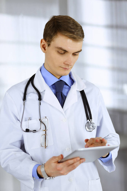 Young doctor man standing at his hospital office, with a stethoscope, using a computer tablet. Perfect medical service in clinic. Happy future in medicine and healthcare - Photo, Image