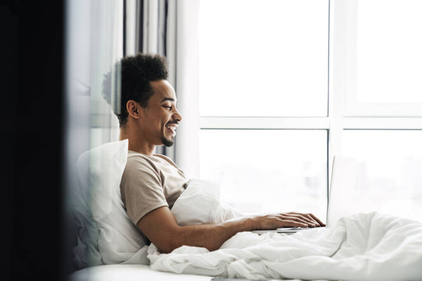 Photo of laughing african american man working with laptop while lying in bed at bright room - Fotó, kép