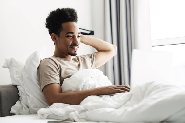 Photo of laughing african american man working with laptop while lying in bed at bright room - Zdjęcie, obraz