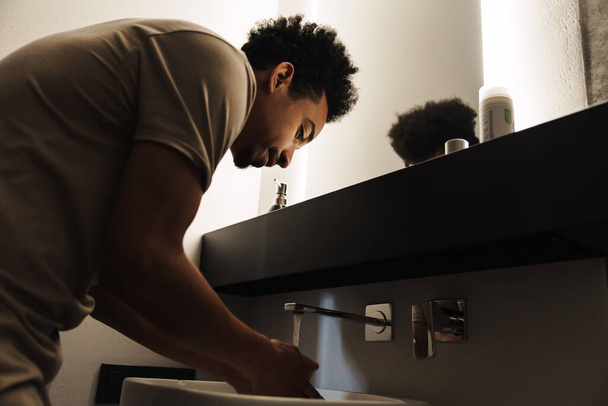 Photo of young african american man with beard cleaning his face in front of mirror at bathroom - Photo, image
