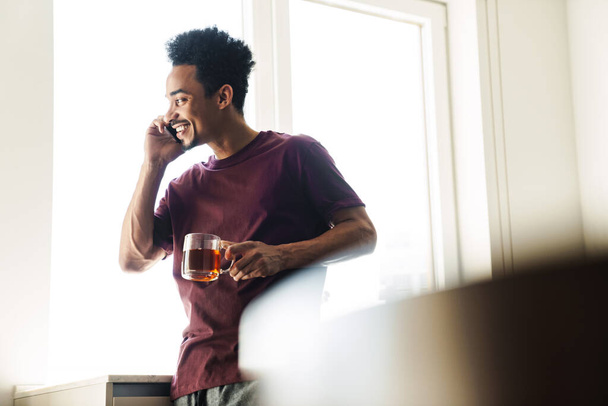 Neşeli Afrikalı Amerikalı bir adamın evde cep telefonuyla çay içerken çekilmiş fotoğrafı. - Fotoğraf, Görsel