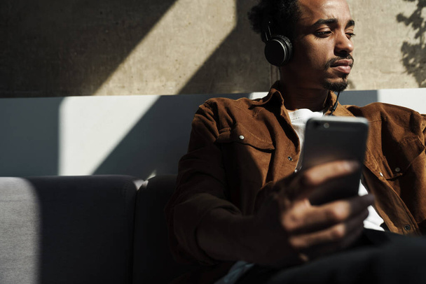 Photo of focused african american man using headphones and cellphone while sitting on sofa in living room - Zdjęcie, obraz