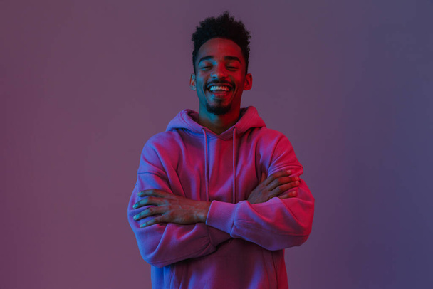 Portrait of smiling african american man in colorful hoodie poising on camera with his arms crossed isolated over violet background - Photo, Image