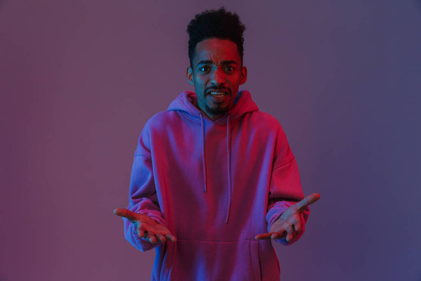 Portrait of irritated african american man in colorful hoodie poising and looking at camera isolated over violet background - Zdjęcie, obraz