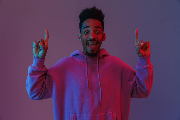 Portrait of exciting african american man in colorful hoodie looking at camera and pointing fingers upward isolated over violet background - Valokuva, kuva